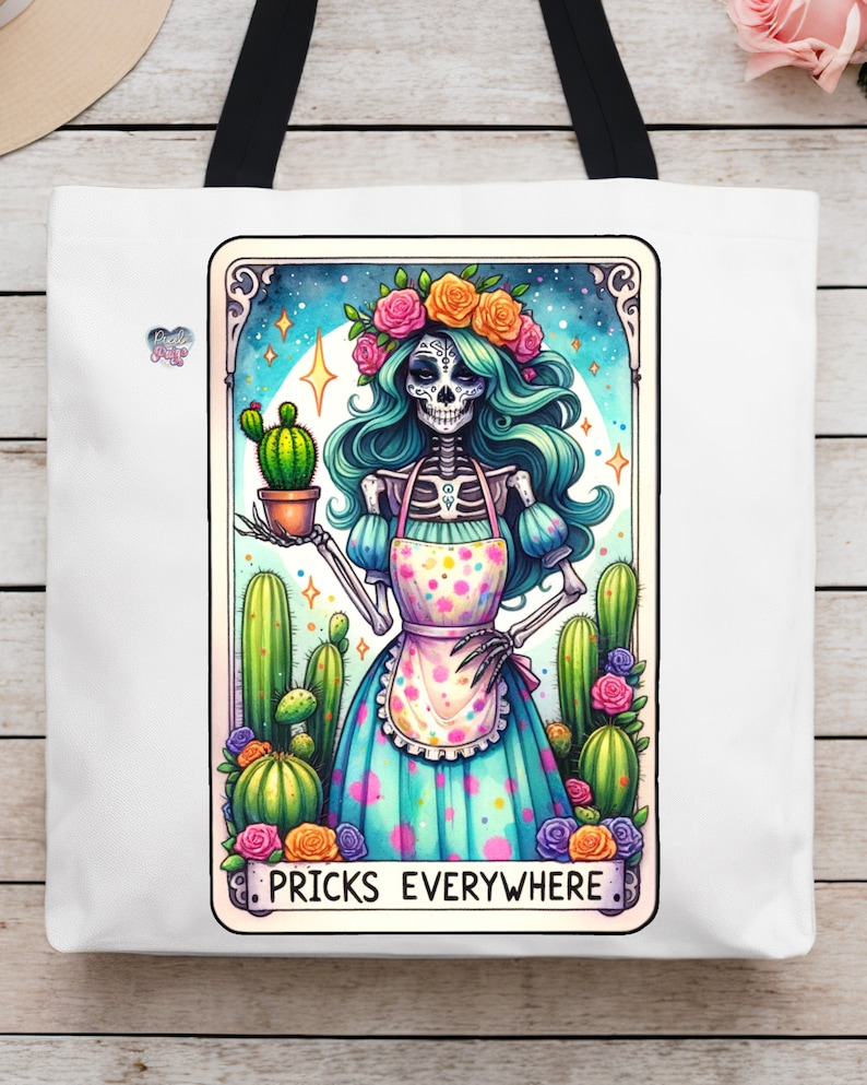 Puede incluir: Una bolsa de tela blanca con una ilustraci&oacute;n colorida de un esqueleto con una corona de flores y un vestido azul y blanco sosteniendo un cactus. El texto "Pricks Everywhere" est&aacute; impreso debajo de la imagen.