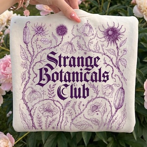 以下が含まれることがあります： 紫色の植物デザインと「Strange Botanicals Club」の文字が入ったクリーム色のスウェットシャツ。 デザインは、さまざまな花のイラストとつる植物が特徴です。 スウェットシャツは、緑の葉とピンクの花を背景にしています。