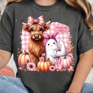 Può includere: T-shirt grigio scuro con un design stravagante. Presenta una mucca delle Highlands con un fiocco floreale, un fantasma che si flette e zucche. La nuvoletta di testo recita "Stronger Than Scary...". Il design include accenti floreali rosa e arancioni.