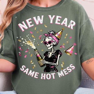 Op de afbeelding: Saliegroen t-shirt met de tekst "NEW YEAR SAME HOT MESS". De afbeelding toont een skelet met een zonnebril, een roze haarband en een feesthoedje, die een champagnefles vasthoudt. Roze confetti en feesthoedjes zijn rond het skelet verspreid.