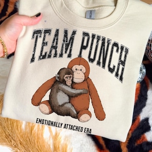 May include: Cream-colored sweatshirt with the text "TEAM PUNCH" in distressed black lettering. Below the text is a graphic of two cartoon monkeys hugging. The phrase "EMOTIONALLY ATTACHED ERA" is printed below the graphic.