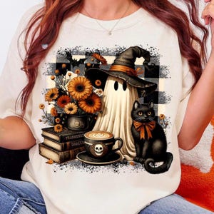 May include: Cream-colored sweatshirt featuring a Halloween-themed graphic. The design includes a ghost wearing a witch hat, a black cat with an orange bow, a coffee cup with a skull, and a stack of books. A bouquet of orange flowers is also present.