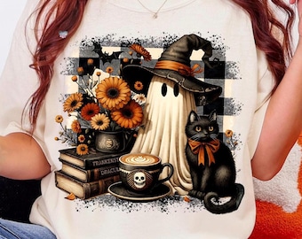 May include: Cream-colored sweatshirt featuring a Halloween-themed graphic. The design includes a ghost wearing a witch hat, a black cat with an orange bow, a coffee cup with a skull, and a stack of books. A bouquet of orange flowers is also present.