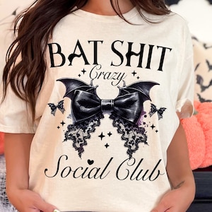May include: A white t-shirt with a black graphic design that says "Bat Shit Crazy Social Club". The graphic features a black bow with bat wings and lace details.