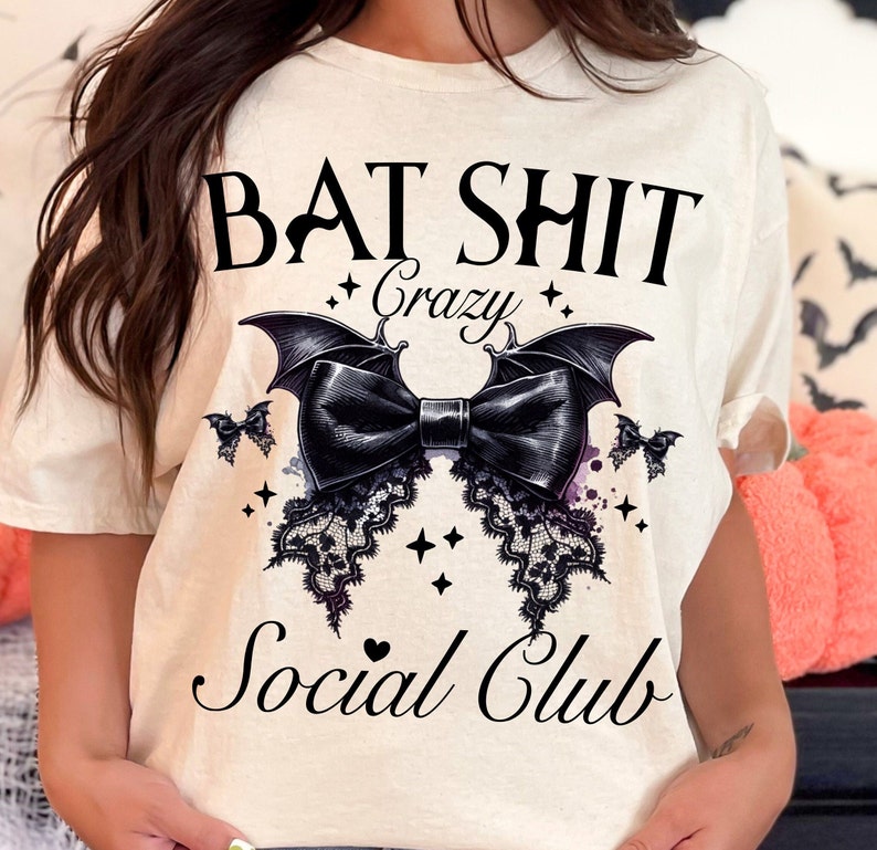 May include: A white t-shirt with a black graphic design that says "Bat Shit Crazy Social Club". The graphic features a black bow with bat wings and lace details.
