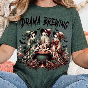 Pode incluir: T-shirt verde com o texto "DRAMA BREWING" acima de um gráfico de três figuras de fantasmas usando chapéus e laços, reunidas em torno de um caldeirão borbulhante. Morcegos e rosas completam o design de Halloween.