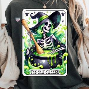 Op de afbeelding: Een tarotkaart-illustratie met een skelet-heks die een groene hoed draagt en een ketel met groene vloeistof roert. De kaart heeft de titel "The Shit Stirrer".