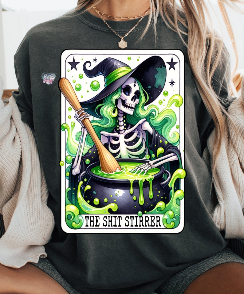 Op de afbeelding: Een tarotkaart-illustratie met een skelet-heks die een groene hoed draagt en een ketel met groene vloeistof roert. De kaart heeft de titel "The Shit Stirrer".