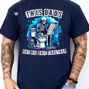 May include: A navy blue t-shirt with a skeleton sitting on a toilet, reading a newspaper and holding a phone. The text on the shirt reads "This Dad's Got His Shit Together".