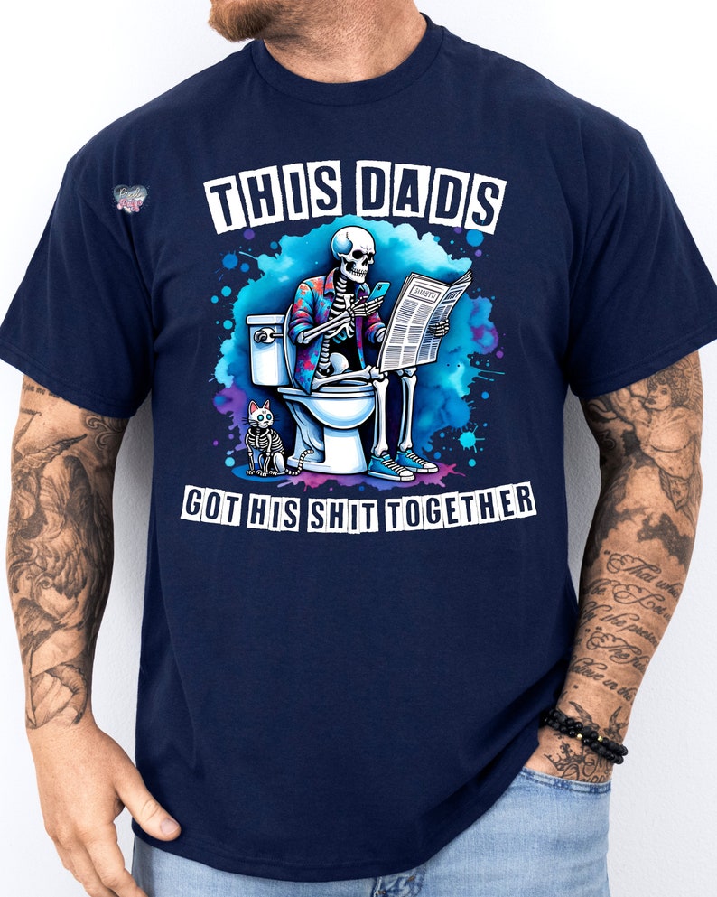 May include: A navy blue t-shirt with a skeleton sitting on a toilet, reading a newspaper and holding a phone. The text on the shirt reads "This Dad's Got His Shit Together".