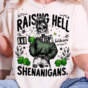 May include: Cream-colored t-shirt with a graphic featuring a skeleton woman holding a green beer. The text reads "RAISING HELL AND SHENANIGANS" with "bad decisions est march 17". Green shamrocks and foliage surround the central image.
