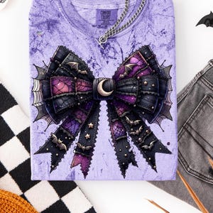 May include: A purple Comfort Colors t-shirt featuring a large, detailed bow design. The bow is black and purple with bat and star accents, and a crescent moon. A silver chain necklace is attached to the shirt. The shirt is folded on a white surface.