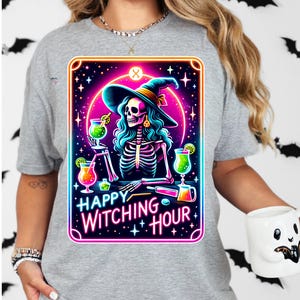 Pu&ograve; includere: Una maglietta grigia con una grafica colorata di una strega scheletro che indossa un cappello da strega e tiene un cocktail. La grafica &egrave; circondata da una cornice rosa e blu al neon e il testo "Happy Witching Hour" &egrave; scritto sotto la grafica.
