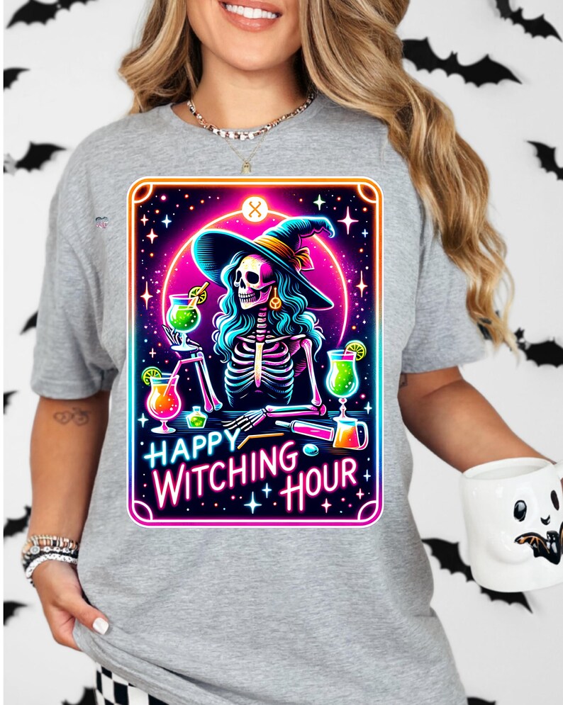 Pu&ograve; includere: Una maglietta grigia con una grafica colorata di una strega scheletro che indossa un cappello da strega e tiene un cocktail. La grafica &egrave; circondata da una cornice rosa e blu al neon e il testo "Happy Witching Hour" &egrave; scritto sotto la grafica.