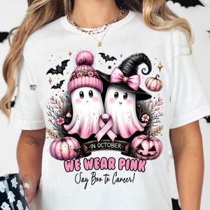Puede incluir: Camiseta blanca con una cinta rosa, dos fantasmas con sombreros de bruja y calabazas. El texto dice "En octubre, usamos rosa, &iexcl;Digamos Boo al c&aacute;ncer!"