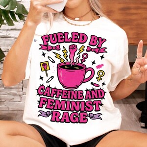 May include: White t-shirt with a pink graphic design that says "Fueled by Caffeine and Feminist Rage". The graphic includes a pink coffee cup with steam coming out of it, a raised fist, and lightning bolts.
