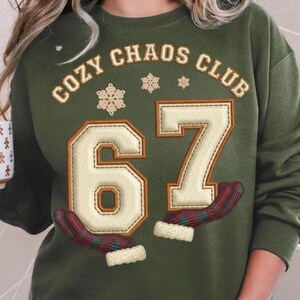 May include: Olive green sweatshirt with the text "COZY CHAOS CLUB" and the number "67" in a cream-colored, stitched design. The number is flanked by two mittens and snowflakes.