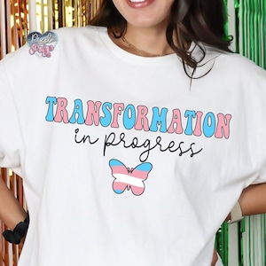 Puede incluir: Camiseta blanca con el texto "TRANSFORMATION in progress" en letras rosa y azul. Debajo del texto hay una mariposa con los colores de la bandera del orgullo transgénero.