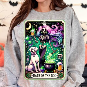 May include: A gray sweatshirt with a graphic print of a skeleton woman with colorful hair holding a beer mug and a paintbrush. The skeleton woman is standing over a cauldron with green smoke coming out of it. A white dog is sitting next to the cauldron. The graphic is surrounded by a green border and the text "Hair of the Dog" is written below the graphic.