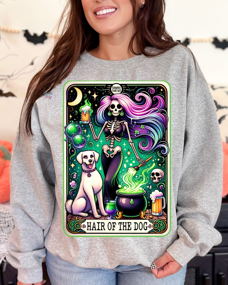 May include: A gray sweatshirt with a graphic print of a skeleton woman with colorful hair holding a beer mug and a paintbrush. The skeleton woman is standing over a cauldron with green smoke coming out of it. A white dog is sitting next to the cauldron. The graphic is surrounded by a green border and the text "Hair of the Dog" is written below the graphic.