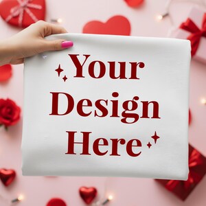 Könnte beinhalten: Ein weißes Tuch mit dem Text "Your Design Here" in fetter, roter Schrift. Das Tuch wird von einer Hand mit pinkfarbenem Nagellack gehalten. Der Hintergrund zeigt rote Herzen, Rosenblätter und eine Geschenkbox.