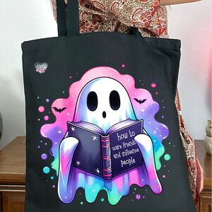 May include: A black tote bag with a ghost holding a book that says "how to scare friends and influence people". The ghost is surrounded by a pink, blue, and green glow.