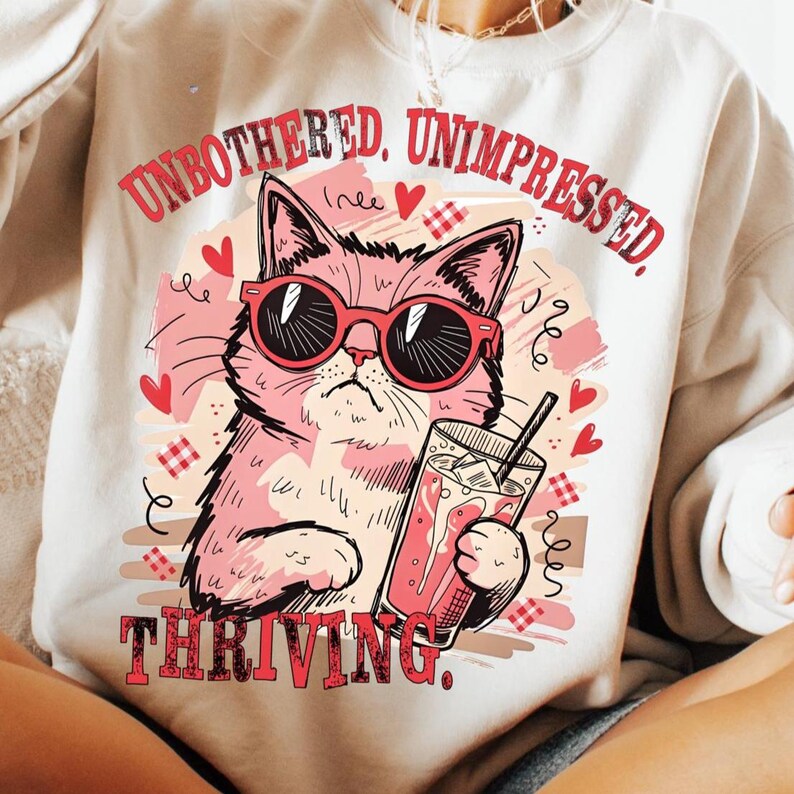 May include: Cream-colored sweatshirt with a pink cat wearing sunglasses, holding a drink with ice and a straw. The text "UNBOTHERED. UNIMPRESSED. THRIVING." is printed in red. Hearts and decorative elements surround the cat.