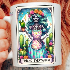 Puede incluir: Taza de cer&aacute;mica blanca con una ilustraci&oacute;n colorida de un esqueleto con una corona de flores y un vestido azul con lunares blancos. El esqueleto sostiene un cactus en maceta y el texto "Pricks Everywhere" est&aacute; impreso debajo de la imagen.