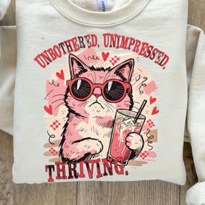 May include: Cream-colored sweatshirt with a pink cat wearing sunglasses and holding a drink. The text "UNBOTHERED, UNIMPRESSED, THRIVING" is printed in red. The design includes hearts and decorative elements.