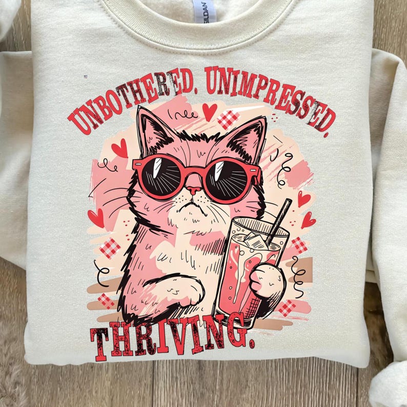 May include: Cream-colored sweatshirt with a pink cat wearing sunglasses and holding a drink. The text "UNBOTHERED, UNIMPRESSED, THRIVING" is printed in red. The design includes hearts and decorative elements.