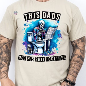 May include: A beige t-shirt with a graphic of a skeleton sitting on a toilet, reading a newspaper and holding a phone. The skeleton is wearing a floral shirt and has a cat sitting next to it. The text on the shirt reads "This Dads Got His Shit Together".