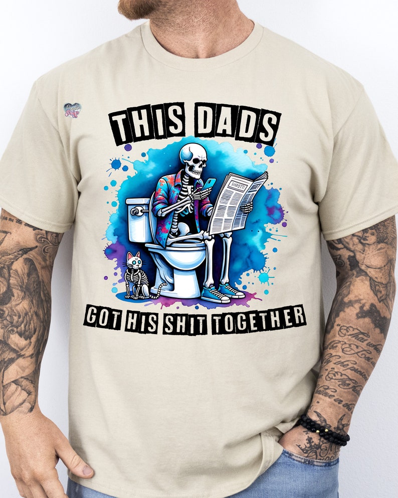May include: A beige t-shirt with a graphic of a skeleton sitting on a toilet, reading a newspaper and holding a phone. The skeleton is wearing a floral shirt and has a cat sitting next to it. The text on the shirt reads "This Dads Got His Shit Together".