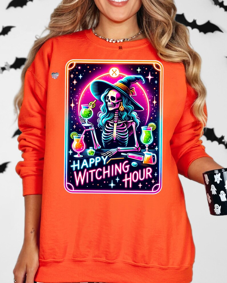 Pu&ograve; includere: Felpa arancione con una grafica di una strega scheletro che indossa un cappello da strega e tiene un cocktail. La grafica &egrave; in stile neon con il testo "Happy Witching Hour".