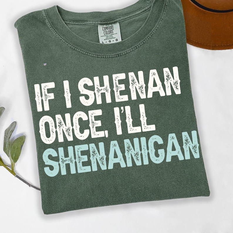 Puede incluir: Una camiseta verde oscuro con el texto "IF I SHENAN ONCE I'LL SHENANIGAN" en letras blancas y celeste. La camiseta est&aacute; doblada y tiene una etiqueta Comfort Colors.