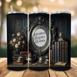 May include: A black and gold stainless steel tumbler with a vintage-inspired design featuring a framed mirror with the text "Dearest Smut Reader", a bouquet of flowers, books, and teacups.
