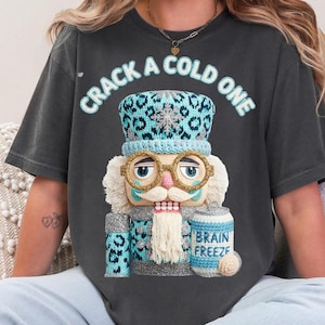 Op de afbeelding: Donkergrijs t-shirt met de tekst "CRACK A COLD ONE" in lichtblauw. Het shirt toont een gedetailleerde illustratie van een notenkraker met een blauw en grijs patroon hoed, bril en sjaal, met een blikje met de tekst "BRAIN FREEZE".