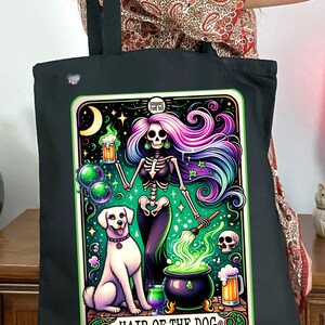 May include: A black tote bag with a colorful illustration of a skeleton witch stirring a cauldron with a dog by her side. The illustration is surrounded by a green border and the text "Hair of the Dog" is at the bottom. The text "Hangover Free Kit" is at the top of the illustration.