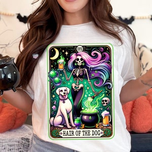 May include: A white t-shirt with a colorful illustration of a skeleton holding a beer mug and a paintbrush, with a white dog sitting next to a cauldron. The text "Hair of the Dog" is written below the illustration.