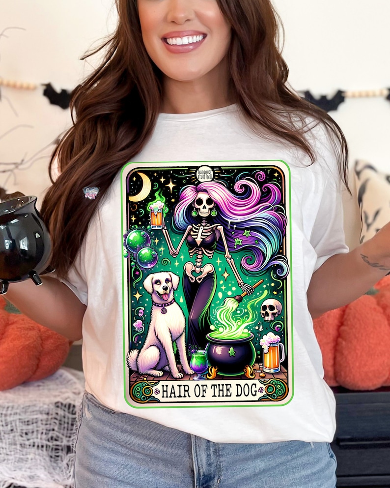 May include: A white t-shirt with a colorful illustration of a skeleton holding a beer mug and a paintbrush, with a white dog sitting next to a cauldron. The text "Hair of the Dog" is written below the illustration.
