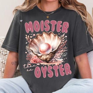 May include: A dark gray t-shirt with a pink graphic of an oyster shell with a pearl inside. The text "MOISTER than an OYSTER" is printed in pink on the shirt.