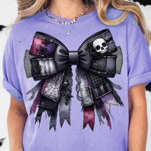 May include: A purple t-shirt with a large bow design. The bow is predominantly black with grey, purple, and pink accents. It features a skull, spiderwebs, and butterfly details, creating a gothic aesthetic.