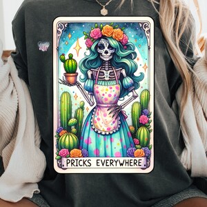Puede incluir: Una ilustraci&oacute;n en blanco y negro de un esqueleto con una corona de flores y un vestido azul y blanco sosteniendo un cactus en maceta. El texto "Pricks Everywhere" est&aacute; debajo de la imagen.