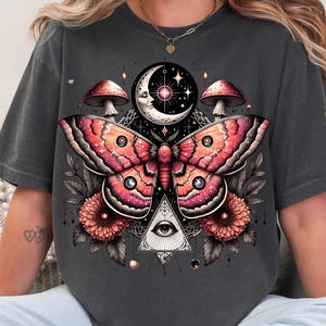 May include: A dark gray t-shirt with a graphic print of a pink and red moth with a crescent moon, stars, mushrooms, and a triangle with an eye in the center.