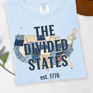 May include: Pale blue t-shirt featuring the text "THE DIVIDED STATES est. 1776" and a patchwork map of the United States. The shirt is made of a soft fabric and has a round neck.