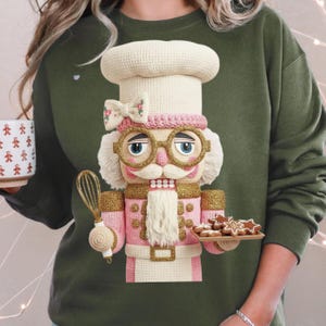 May include: Green sweatshirt featuring a detailed, crocheted nutcracker chef design. The nutcracker wears a white chef's hat, pink jacket, and gold-rimmed glasses. It holds a whisk and a tray of gingerbread cookies, perfect for the holiday season.