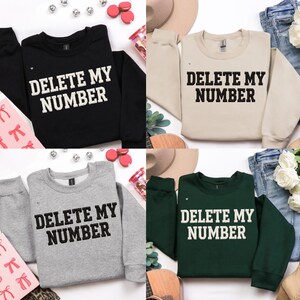May include: Four sweatshirts in black, beige, gray, and dark green, each with the text "DELETE MY NUMBER" in bold white letters. The sweatshirts are displayed with accessories like bows, hats, and jeans.
