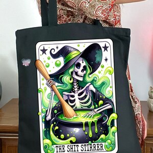 Op de afbeelding: Zwarte tote bag met een groene en witte illustratie van een skelet-heks die een ketel roert met een houten lepel. De tekst "The Shit Stirrer" is onder de afbeelding gedrukt.
