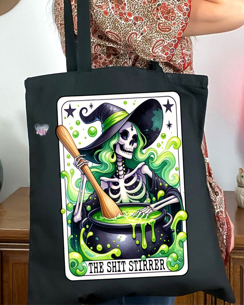 Op de afbeelding: Zwarte tote bag met een groene en witte illustratie van een skelet-heks die een ketel roert met een houten lepel. De tekst "The Shit Stirrer" is onder de afbeelding gedrukt.