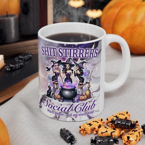 Op de afbeelding: Witte keramische mok met een paars, zwart en wit ontwerp met drie heksen die een ketel roeren. De tekst "Shit Stirrers Social Club" is op de mok gedrukt. De tekst "Witch please...tell me everything!" is onder het ontwerp gedrukt.