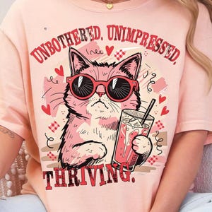 May include: A light pink t-shirt with a graphic of a pink cat wearing red sunglasses and holding a drink. The text "UNBOTHERED, UNIMPRESSED, THRIVING" is printed above and below the cat illustration.